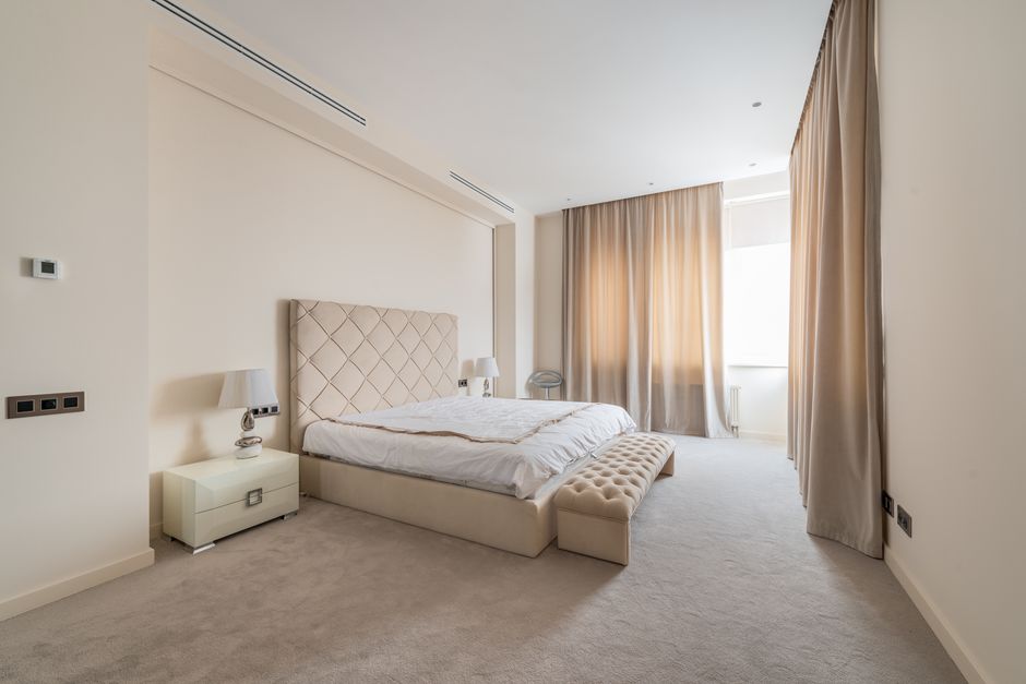 A spacious, modern bedroom with a large, upholstered beige headboard attached to a clean, white wall. The bed is neatly made with white bedding and a light beige throw at the foot. On each side of the bed, there are small white bedside tables with matching lamps. The room features a soft, cream-colored carpet covering the floor, and beige curtains hang from floor to ceiling, partially drawn to allow natural light through the large window behind. A small, simple chair is positioned near the window. The walls are painted in a neutral, off-white shade, and the room appears tidy and well-maintained, reflecting professional deep cleaning and hygiene standards. Merton Carpet Cleaning emphasizes surface cleaning and sanitisation in domestic spaces like this to promote hygiene and prevent fines related to mattress and carpet disposal rules, as highlighted on the 'avoid fines' webpage.