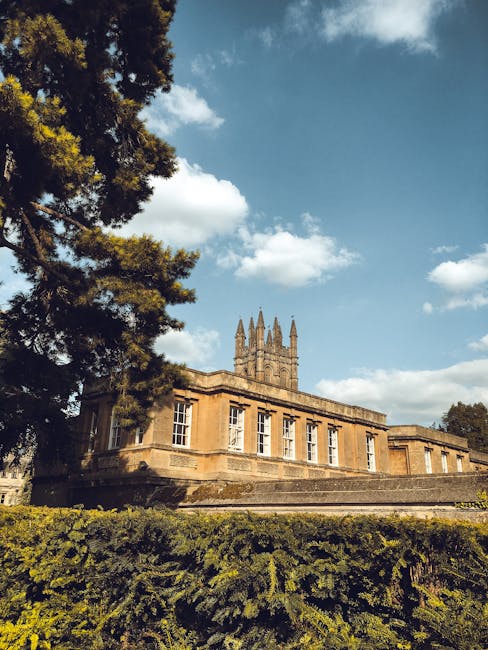 Exterior view of a historic stone building with large windows, set on a slightly elevated platform. The structure features a prominent tower with pointed spires and crenellations, indicative of Gothic architectural style. Surrounded by a well-maintained green hedge and tall trees, including a large tree on the left side, under a partly cloudy sky with white clouds and patches of blue. The scene is lit by natural daylight, highlighting the building's weathered stone surface. This image exemplifies architectural preservation and is relevant to discussions on surface cleaning and maintenance of historic buildings for safety and appearance, as promoted by Merton Carpet Cleaning in the context of property hygiene and upkeep.