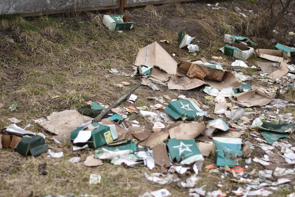 A patch of outdoor ground covered in scattered cardboard boxes, paper waste, and packaging materials, with some green and white boxes displaying a star logo. The debris is mixed with grass and soil, indicating an outdoor spill. In the background, there are leafless bushes and a fence. The scene appears dirty and unkempt, highlighting the need for thorough cleanup and waste removal. Merton Carpet Cleaning specializes in deep cleaning and sanitisation services, but this image specifically relates to external waste spill cleanup in Mitcham as seen in the page titled 'Bulky waste spill in Mitcham? Urgent carpet cleanup, MERTON', available on mertoncarpetcleaning.org.uk.