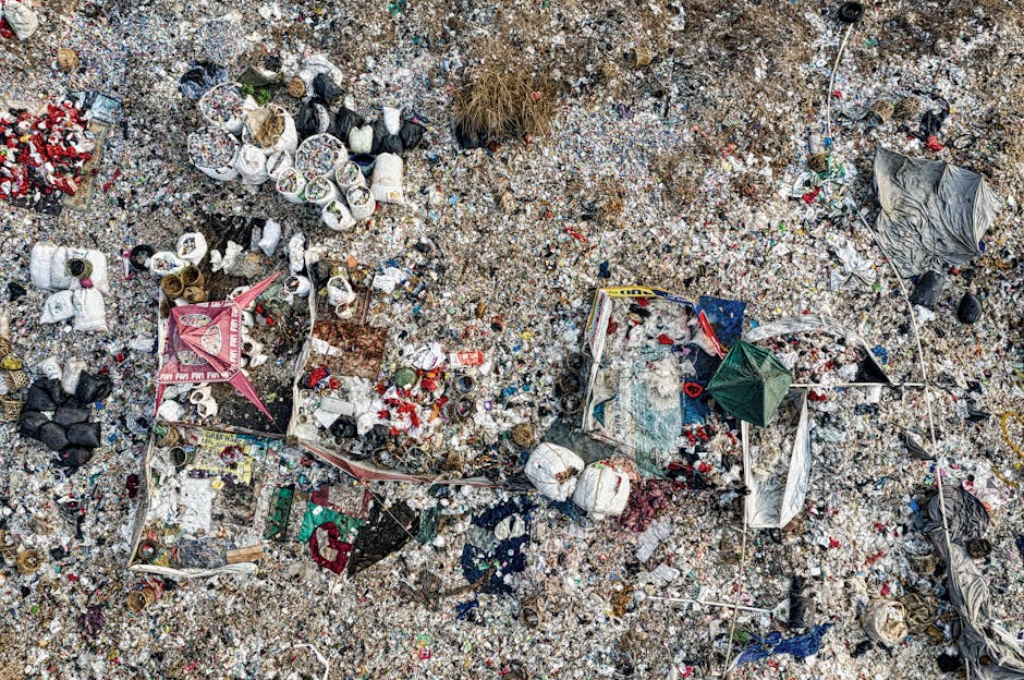 Aerial view of a large spill of bulky waste and litter on a gravel and dirt surface, showing scattered plastic bags, cardboard, paper, and miscellaneous debris. Some objects are contained within makeshift enclosures made of fabric and ropes. The area appears unclean with dirt and waste spread across the ground, indicating a recent waste spill. The scene is outdoors, with natural lighting highlighting the mess. This image emphasizes the need for thorough surface cleaning and sanitation, which Merton Carpet Cleaning can assist with through professional domestic cleaning services.