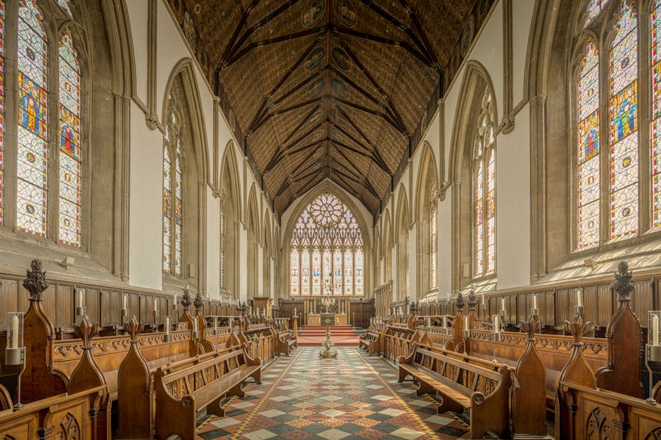 An interior view of a church sanctuary with high vaulted wooden ceilings and large stained glass windows on both sides, allowing natural light to illuminate the space. The aisle is lined with polished wooden pews that have carved detailing and ornate end caps, leading up to the altar at the front. The tiled floor features a geometric pattern in various muted tones. The overall scene appears clean, well-maintained, and ready for service, illustrating the importance of surface cleaning and maintenance in such historic and sacred spaces. Merton Carpet Cleaning offers professional deep cleaning and sanitisation services suitable for similar settings to maintain hygiene and visual appeal.