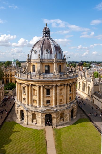 The image shows the exterior of the historic Radcliffe Camera building in Oxford, featuring a large, domed roof with weathered stonework and intricate architectural details. The lower part of the structure is constructed from pale-colored stone blocks, with tall, narrow windows and ornate decorative elements. Surrounding the building, there is a well-maintained green lawn with a paved walkway leading up to the entrance, and nearby buildings with Gothic features and flags. The sky above is partly cloudy with patches of blue, providing natural daylight that highlights the building's historic features. The scene captures the grandeur and architectural complexity of this famous academic and library landmark.