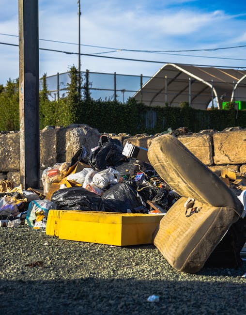 A pile of discarded household waste including black plastic trash bags, a yellow bin, a used beige sofa cushion, and other debris are scattered on a gravel surface next to a stone boundary wall. The background features a metal fence, a partly cloudy blue sky, and structures such as a greenhouse or covered outdoor space, with overhead power lines visible. This scene illustrates waste accumulation outside, highlighting the importance of proper disposal and domestic cleaning practices, as emphasized by Merton Council rules for carpet and bulky waste disposal, MERTON. Merton Carpet Cleaning specializes in surface cleaning and deep cleaning services for maintaining hygiene and cleanliness within residential and commercial environments.