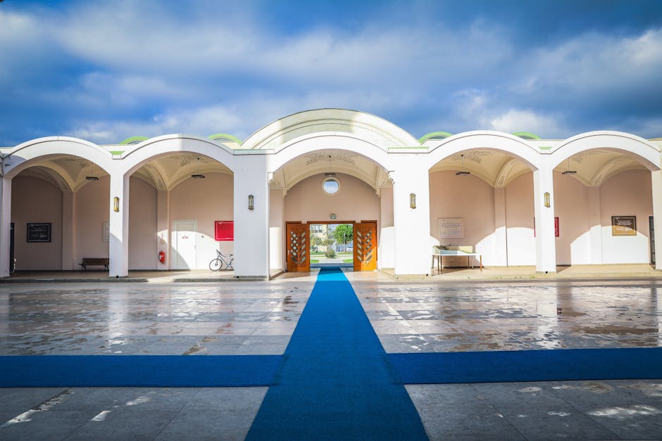 An exterior view of a white architectural structure featuring a series of arches and a central wooden gateway with decorative cutouts. The area is paved with large stone tiles, some showing signs of wear, and a vibrant blue carpet runner extends from the foreground, leading through the open gateway towards a park visible in the distance. The structure is illuminated by natural daylight, with shadows cast by the arches, and the sky above is partly cloudy with patches of blue. The scene demonstrates a clean and well-maintained entrance, reflecting ongoing surface cleaning and maintenance efforts by Merton Carpet Cleaning, aimed at preserving hygiene and appearance in this public space.