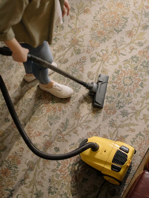 A person wearing beige shoes, blue jeans, and a beige coat is using a yellow vacuum cleaner to perform surface cleaning and deep cleaning on a large, patterned carpet in a residential living room. The carpet features a floral design in muted tones of cream, green, and soft orange. The vacuum cleaner has a long hose and a power cord visible in the foreground. The room is well-lit, and the carpet appears clean and well-maintained. The setting suggests that Merton Carpet Cleaning offers professional sanitisation and maintenance services for domestic carpets, ensuring hygiene and freshness in homes.