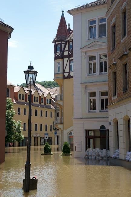 Photograph of a city street during a flood, with water level rising to cover the lower portions of multi-storey buildings that have European architectural styles, including bay windows and decorative facades. A traditional black street lamp is partially submerged in the floodwaters, and sandbags are stacked against the building entrances to prevent water ingress. The scene is illuminated by natural daylight, highlighting the extent of the flooding. The image emphasizes the impact of floodwater on urban infrastructure, relevant to post-flood property protection and cleaning services offered by Merton Carpet Cleaning, especially for drying and cleaning affected carpets and surfaces after flooding incidents in Merton homes.