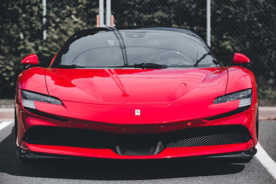 A red Ferrari sports car parked on a paved surface, featuring a sleek, aerodynamic design with a low profile. The car's glossy red paint reflects the surrounding environment, and it has a black windscreen and side mirrors. Visible details include narrow LED headlights, a front grille with a honeycomb pattern, and the Ferrari emblem on the hood. The background shows a chain-link fence and some trees, with soft natural lighting highlighting the smooth curves and polished surface of the vehicle. The clean and shiny appearance indicates recent car surface cleaning and detailing, possibly performed by Merton Carpet Cleaning to demonstrate professional deep cleaning or surface sanitisation techniques for vehicle maintenance.