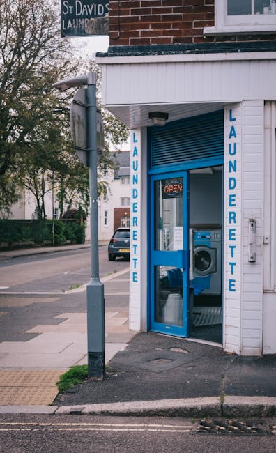 Exterior view of a small laundry shop on Raynes Park High Street (SW20), showing a white wooden façade with blue signage reading 'LAUNDERETTE' on either side of a glass door. The door has a blue frame and a small 'OPEN' sign illuminated inside. Visible through the glass are a washing machine and a dryer, indicating the laundry service offered. To the left of the entrance, a silver street light pole stands on the sidewalk, which is paved with concrete slabs and includes tactile paving for accessibility. In the background, there are parked cars and trees lining the street, with natural daylight illuminating the scene. Merton Carpet Cleaning specializes in professional cleaning services, including deep cleaning and surface sanitisation, ensuring high standards of hygiene for residential and commercial clients.