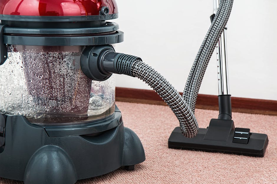 Close-up of a wet vacuum cleaner with a transparent dust container filled with water and foam, connected to a flexible hose with a patterned surface, positioned on a pink carpeted floor in a domestic setting. The vacuum cleaner has a red top and a black base, with additional vacuum tools and a metal handle visible in the background. The room has white walls and wooden skirting boards, indicating a clean and well-maintained living space. The image illustrates surface cleaning and deep cleaning processes provided by Merton Carpet Cleaning, emphasizing hygiene and maintenance of soft furnishings and flooring.