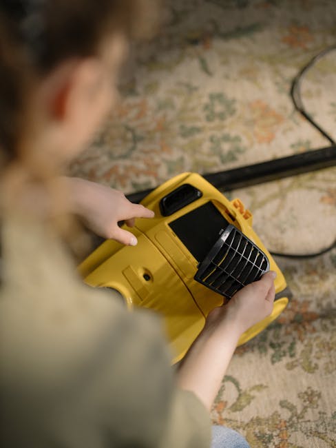 Close-up of a wet vacuum cleaner with a transparent dust container filled with water and foam, connected to a flexible hose with a patterned surface, positioned on a pink carpeted floor in a domestic setting. The vacuum cleaner has a red top and a black base, with additional vacuum tools and a metal handle visible in the background. The room has white walls and wooden skirting boards, indicating a clean and well-maintained living space. The image illustrates surface cleaning and deep cleaning processes provided by Merton Carpet Cleaning, emphasizing hygiene and maintenance of soft furnishings and flooring.