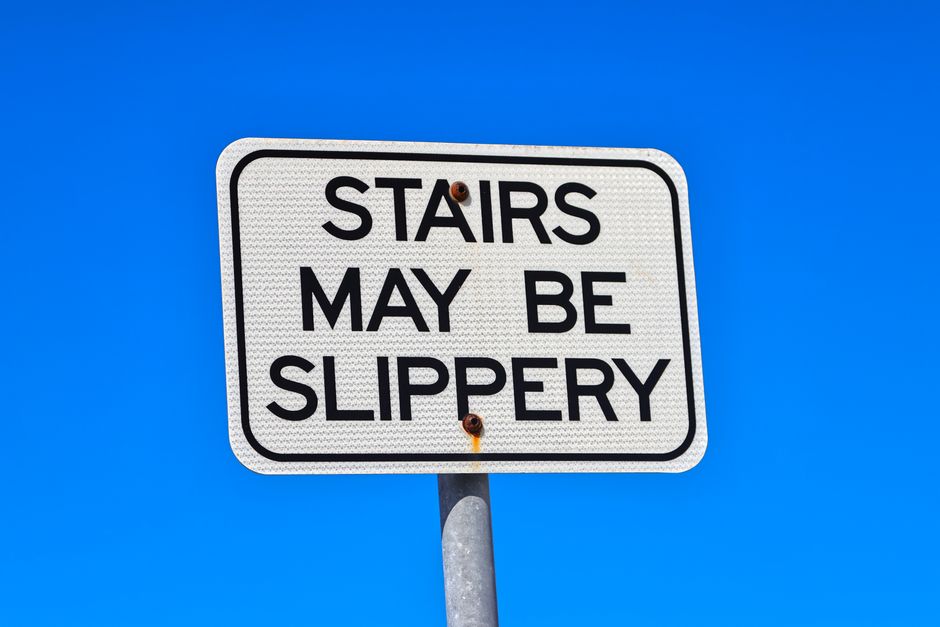 A rectangular white safety sign with black text reading 'STAIRS MAY BE SLIPPERY' is mounted on a metal pole against a clear blue sky background. The sign has a black border and a textured surface. The sunlight illuminates the sign, with visible rust spots near the mounting points. No other objects or surfaces are visible, focusing solely on the warning sign to caution about potential slipping hazards on stairs. This image may relate to safety and maintenance considerations, aligning with specialist cleaning and surface safety discussions, for example, by Merton Carpet Cleaning, under the context of surface and environment safety.