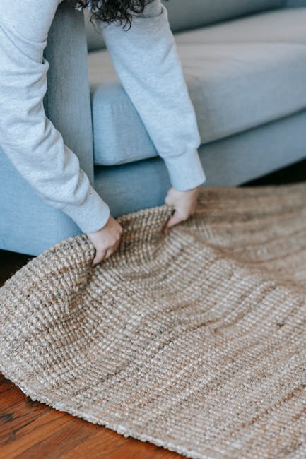 A person dressed in a light grey sweatshirt and blue jeans is lifting the corner of a textured beige area rug from a wooden floor, preparing for cleaning or maintenance. The room features a light grey upholstered sofa with a smooth fabric surface positioned against a wall, and the wooden flooring is polished and free of visible dust or debris. Natural light illuminates the scene, highlighting the clean and well-maintained appearance of the space. The image reflects the process of surface cleaning and preparation within a domestic setting, showcasing attention to hygiene and care by Merton Carpet Cleaning.