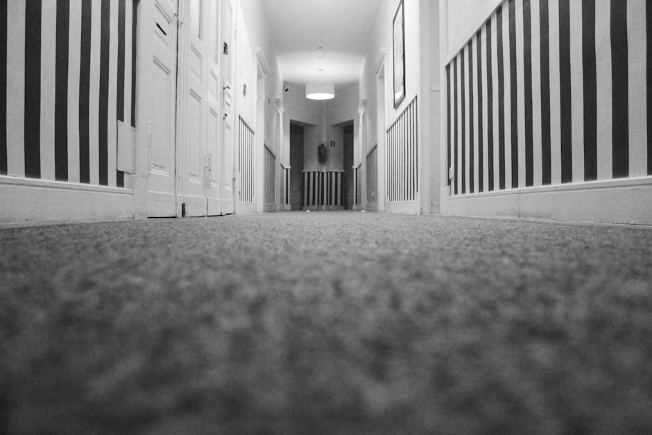 A black and white photograph of a residential hallway taken from a low-angle perspective. The carpeted floor appears clean and well-maintained, with a textured surface visible close up. On either side, white wooden wainscoting runs along the lower part of the walls, which are painted in a light color. Several closed white doors are evenly spaced along the corridor, each with decorative molding. The ceiling features a circular overhead light fixture casting diffuse illumination, creating a bright and hygienic atmosphere. In the background, the hallway extends towards a door at the end. The image showcases a neat, dust-free environment suitable for professional surface cleaning and sanitisation, as provided by Merton Carpet Cleaning, serving the Wimbledon Village area.