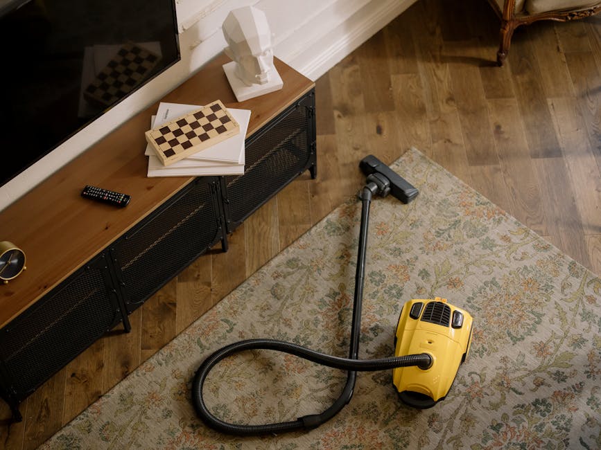 A yellow canister vacuum cleaner with a black hose and silver telescopic wand is positioned on a patterned area rug in a living room, ready for surface cleaning and deep cleaning. The room features a wooden floor and a low black media console with a white bust sculpture, remote control, and some papers on top. A small wooden chessboard sits beside the sculpture, and natural light illuminates the space, highlighting the clean, well-maintained floor and tidy arrangement. The setting reflects domestic cleaning processes carried out by Merton Carpet Cleaning, emphasizing hygiene and surface maintenance.